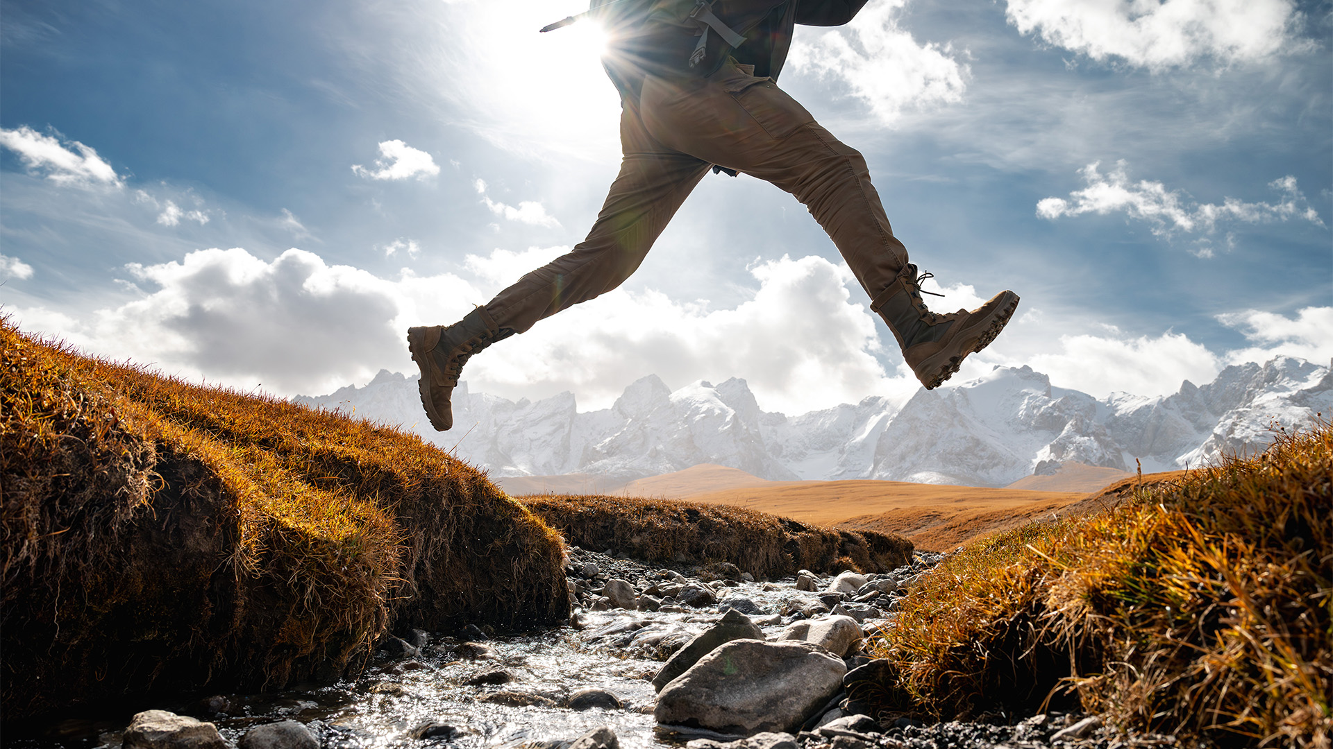 A male hiker leaping over a flowing brook lined by red grass and silhouetted against a blue sky